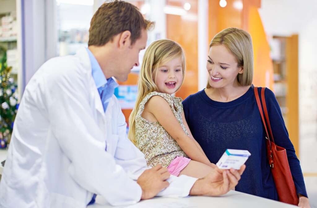 A pharmacist showing a medicine box to a smiling mother and her young daughter at the pharmacy counter